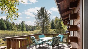 Terraza de madera con mesa y sillas, bordeada por una barandilla de madera, con vistas a un área boscosa.