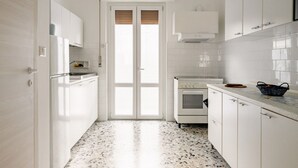 Une cuisine étroite avec des meubles blancs, un réfrigérateur blanc, une cuisinière et une porte dotée d’une fenêtre.