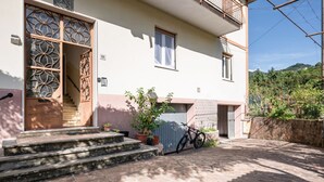 Une maison à deux étages avec un escalier menant à l’entrée, un vélo garé à l’extérieur et une vue sur la verdure environnante.