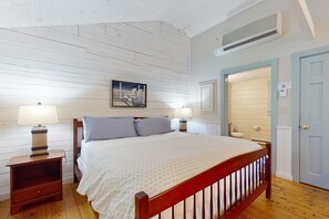 A bedroom with a wooden bed, two bedside tables with lamps, a wall-mounted air conditioner, and a door with a light switch. - Point Lookout (Lincolnville)