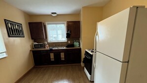 A compact kitchen with wooden flooring, a white refrigerator, dark cabinets, a microwave, and a window with blinds.