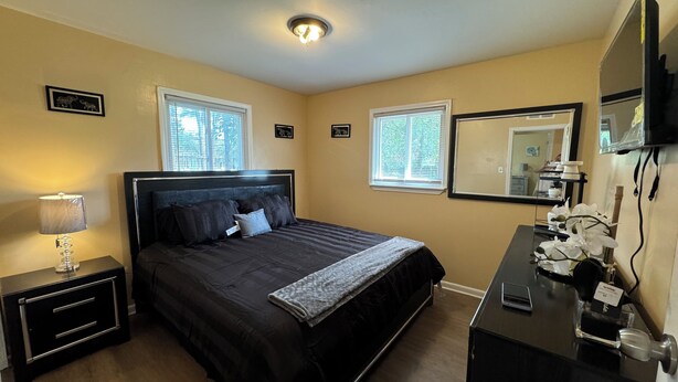 A bedroom with a bed, bedside table, lamp, dresser, mirror, and windows.