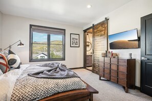 Une chambre avec une grande fenêtre, un lit recouvert d’une couverture à motifs, une commode en bois et un téléviseur fixé au mur.