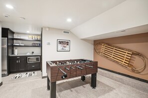 A room with a foosball table, a kitchen area with shelves, and a wall-mounted sled.