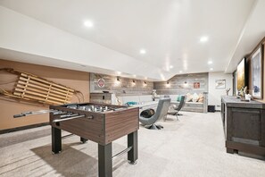 Un espacio interior moderno con mesa de fútbol, zona de estar con sofá y sillones, y pared de madera con estante colgante.