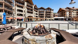 Un foc de pedra amb seients, envoltat per una paret de pedra, en una plaça amb edificis de fusta i zones d'estar a l'aire lliure.