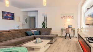 Ein modernes Wohnzimmer mit einem braunen Sofa, einem weißen Couchtisch und einem Schreibtisch mit einem Computer. Ein Kamin mit rotem Licht, ein Bild an der Wand und eine Pflanze in der Ecke sind ebenfalls vorhanden.