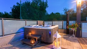 Ein Whirlpool mit einer Holzbank und zwei Kerzen, umgeben von einer Terrasse mit Geländer und einem Blumentopf.