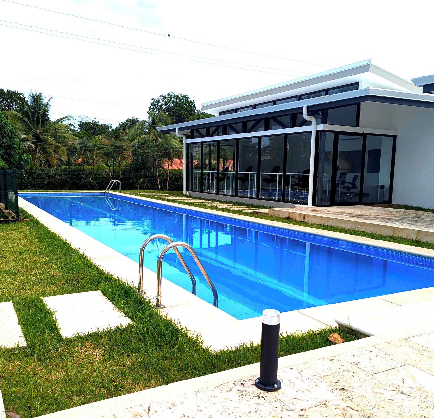 A modern swimming pool with clear blue water, surrounded by a well-maintained lawn and a glass-enclosed building with a contemporary design.