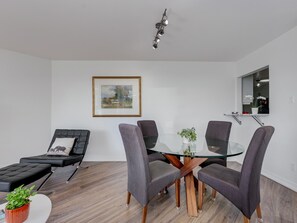 Una sala de estar moderna con una mesa de comedor de vidrio, sillas tapizadas en gris, un sofá negro, un cuadro enmarcado y una planta en maceta.