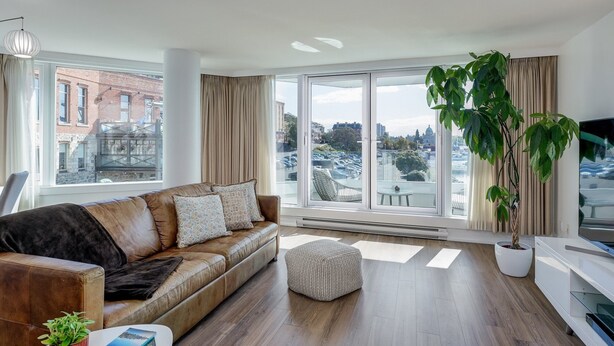 A modern living room with a leather sofa, a small ottoman, a coffee table with a book and a plant, a large window with curtains, and a potted plant. - Coastal Charm – Breathtaking Views with Work Desk by Victoria Prime! (Victoria)