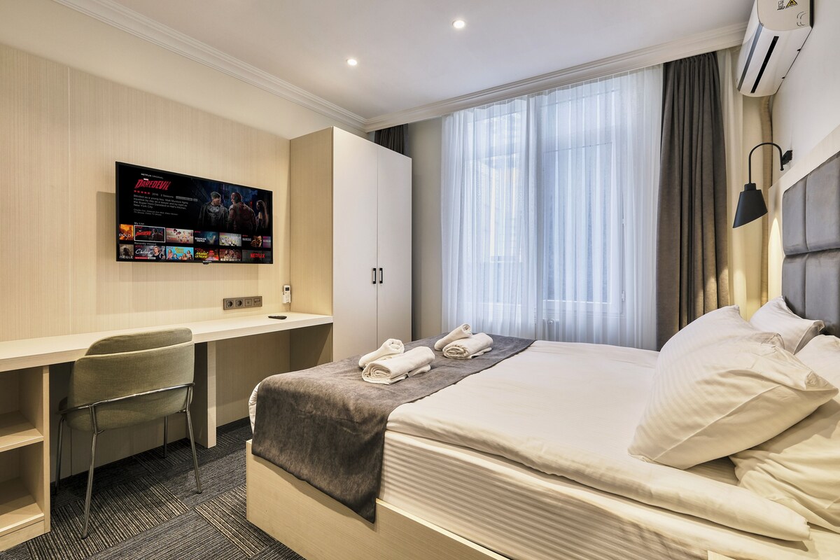 A hotel room with a bed, a desk, a chair, a TV mounted on the wall, and a window with curtains.