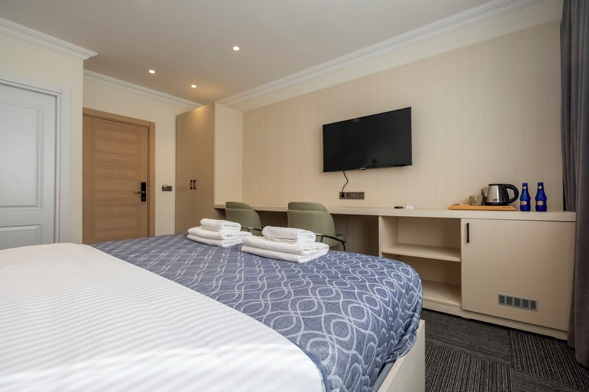 A hotel room with a large bed, a flat-screen TV mounted on the wall, a desk with chairs, and a minibar.