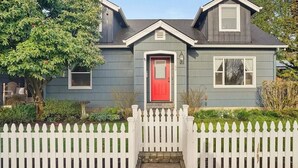 Una casa de dos pisos con una puerta roja, una valla de piquetes blancos y un pequeño jardín delantero.