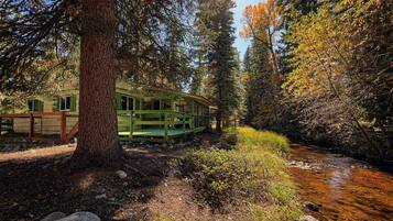 Un chalet rustique doté d’une terrasse en bois, entouré d’une forêt dense et bordé par un ruisseau.