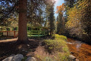 Una cabaña rústica con terraza de madera, rodeada de un bosque denso y un arroyo.