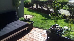 A patio with a black sofa, a small table, and potted plants. There is a view of a road and a red car in the background.