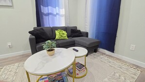 Ein Wohnzimmer mit grauem Sofa, einem kleinen runden Tisch mit Pflanze und einer Fernbedienung.
