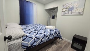 Ein Schlafzimmer mit einem Bett, einer blau-weißen Bettdecke, einem weißen Kopfkissen, einem blauen Vorhang, einem Blumenbild an der Wand und einer kleinen, schwarzen Kommode.