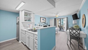 A modern kitchen with white cabinets, a central island, and a dining area with a table and chairs. - Sugar Beach 135: Your Modern Coastal Studio (Orange Beach)