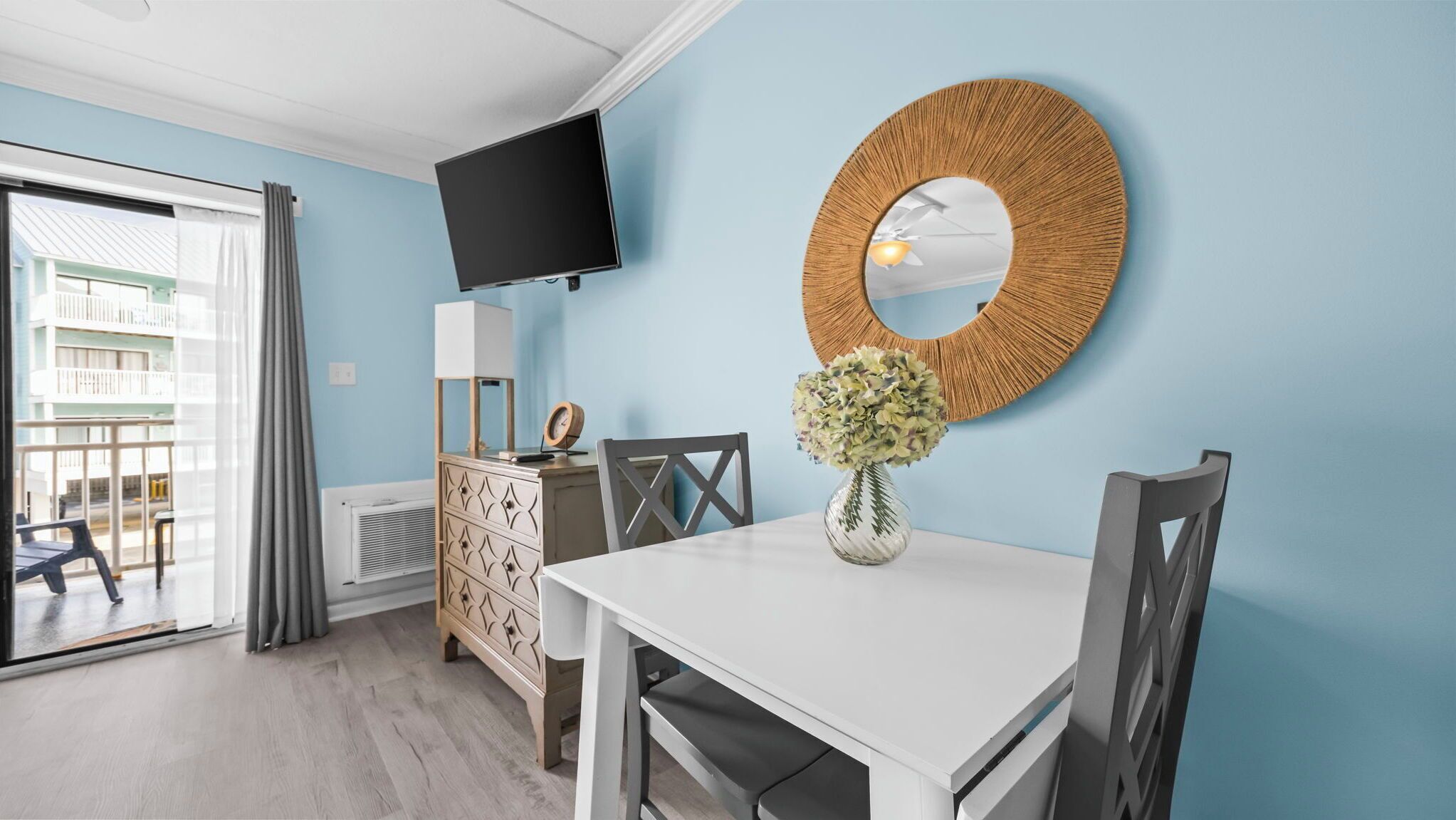 A room with a white table, a round mirror, a vase with flowers, a TV on the wall, and a view of a balcony with outdoor furniture.
