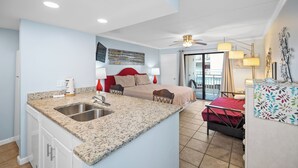 Habitación de hotel compacta con kitchenette, cama con ropa de cama roja, escritorio pequeño y balcón con vistas.