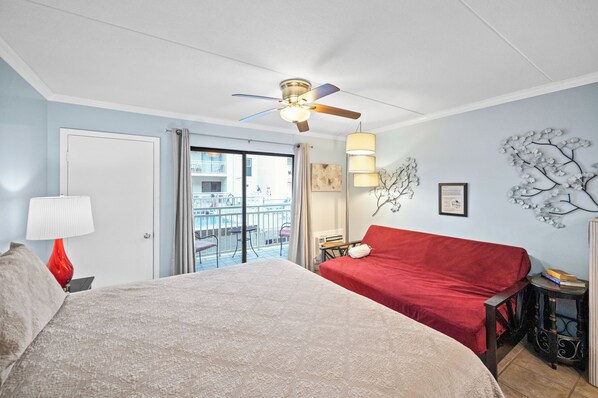 A hotel room with a bed, a red sofa, a ceiling fan, a balcony with a view, and a decorative wall piece. - Sugar Beach 160 Pool View (Orange Beach)