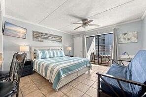 A bedroom with a bed, a chair, a desk, a ceiling fan, and a balcony with a view of a building.