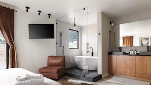 A modern bedroom with a bed, a brown leather chair, a glass-enclosed shower, a white bathtub, and a wooden vanity with a mirror. - Relaxing Wellness Escape with Sauna & Cold Plunge (Austin)