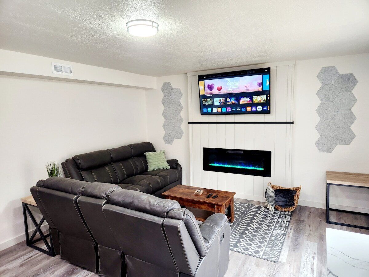 A modern living room with a black leather sofa, a wooden coffee table, a flat-screen TV mounted on the wall, and a patterned rug.