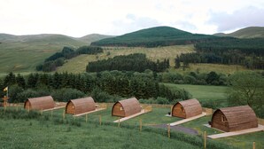 Une rangée de cabanes en bois se dresse dans un champ verdoyant, avec une montagne en toile de fond.