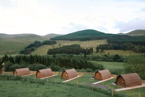 A row of wooden cabins in a grassy field with a mountain backdrop. - Cosy Rural Cabin With Hillside Viewpoint (Moffat)