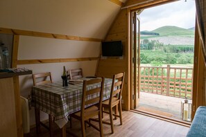 A dining area with a table set for two, wooden chairs, a plaid tablecloth, and a bottle of wine. A television is mounted on the wall, and a view of a forested landscape is visible through an open door. - Cosy Rural Cabin With Hillside Viewpoint (Moffat)