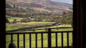 Une vue depuis l’intérieur d’une pièce dont la fenêtre donne sur un paysage rural, avec des collines ondoyantes et une petite maison au loin.