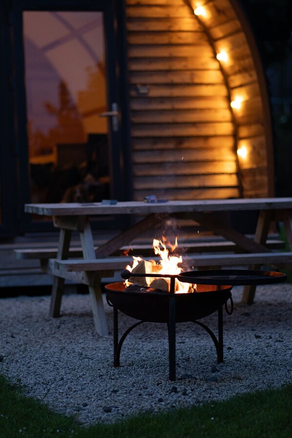 A fire pit with flames, a wooden picnic table, and a building with a curved wooden exterior in the background.