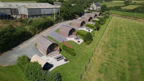 A row of wooden arch-roofed cottages with outdoor seating areas, set in a rural landscape with a large barn and open fields. - Cosy Rural Cabin in the Cornish Countryside (Helston)