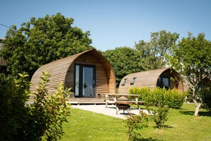 Two wooden cabins with large sliding doors, a picnic table, and a small wheelbarrow in a grassy area with trees. - Cosy Cabin with Hot Tub in the Cornish Countryside (Helston)