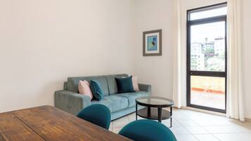 A modern living room with a grey sofa, teal chairs, a wooden table, and a large window with a view of buildings and a balcony.