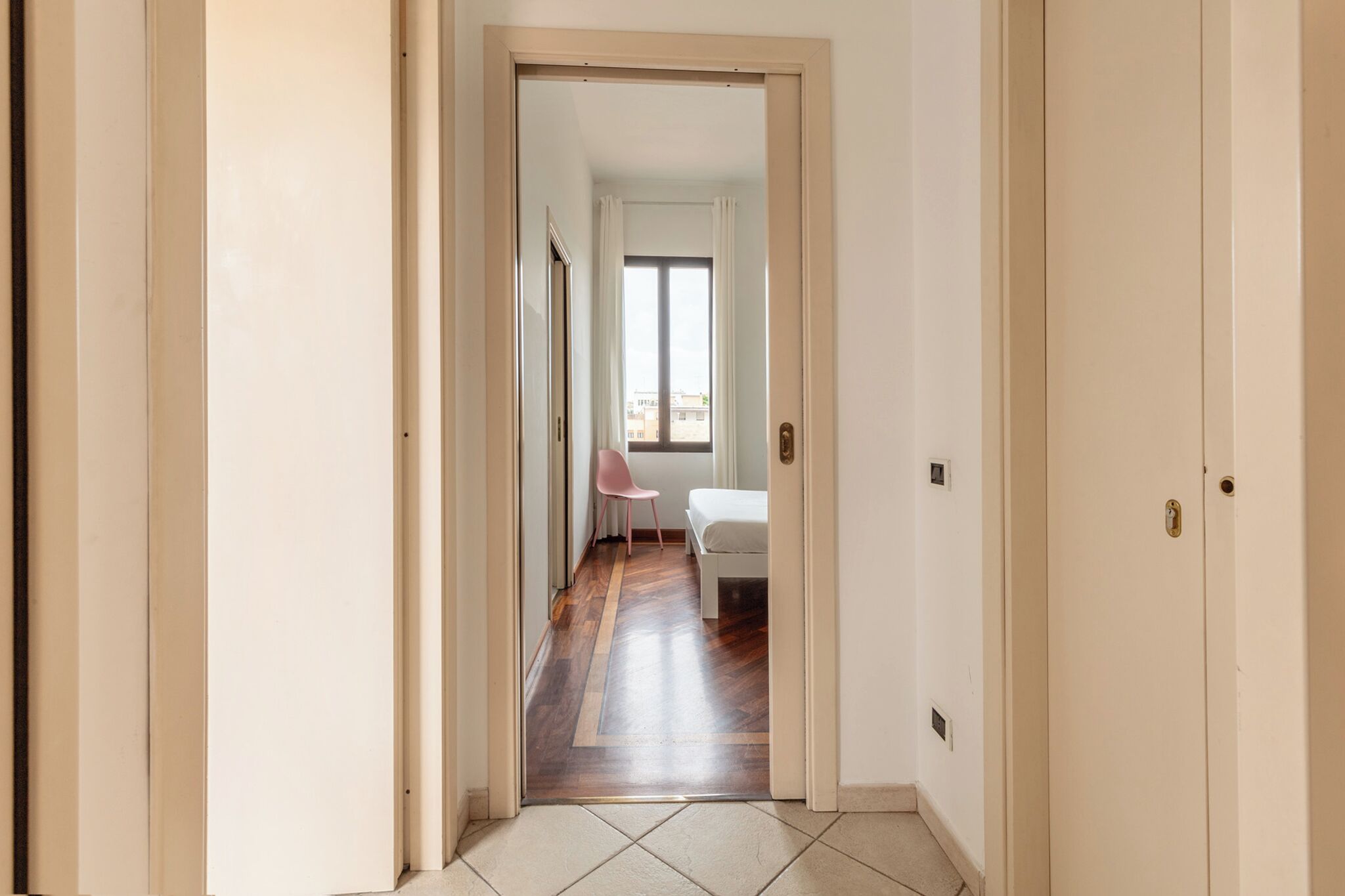 A hallway with a wooden floor, a white door, and a view into a room with a bed and a chair.