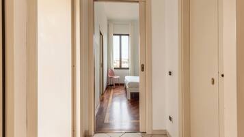 A hallway with a wooden floor, a white door, and a view into a room with a bed and a chair.