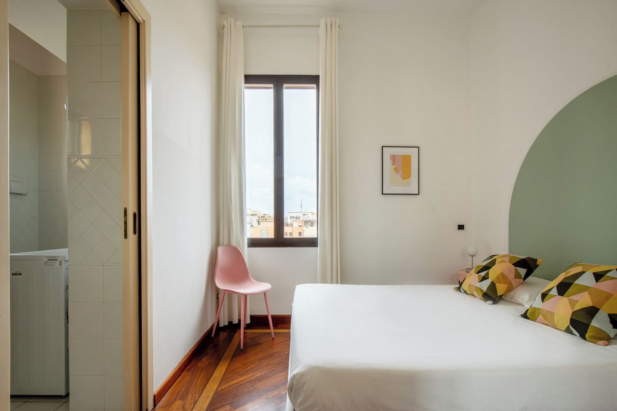 A bedroom with a bed, a chair, a window, and a view of buildings.