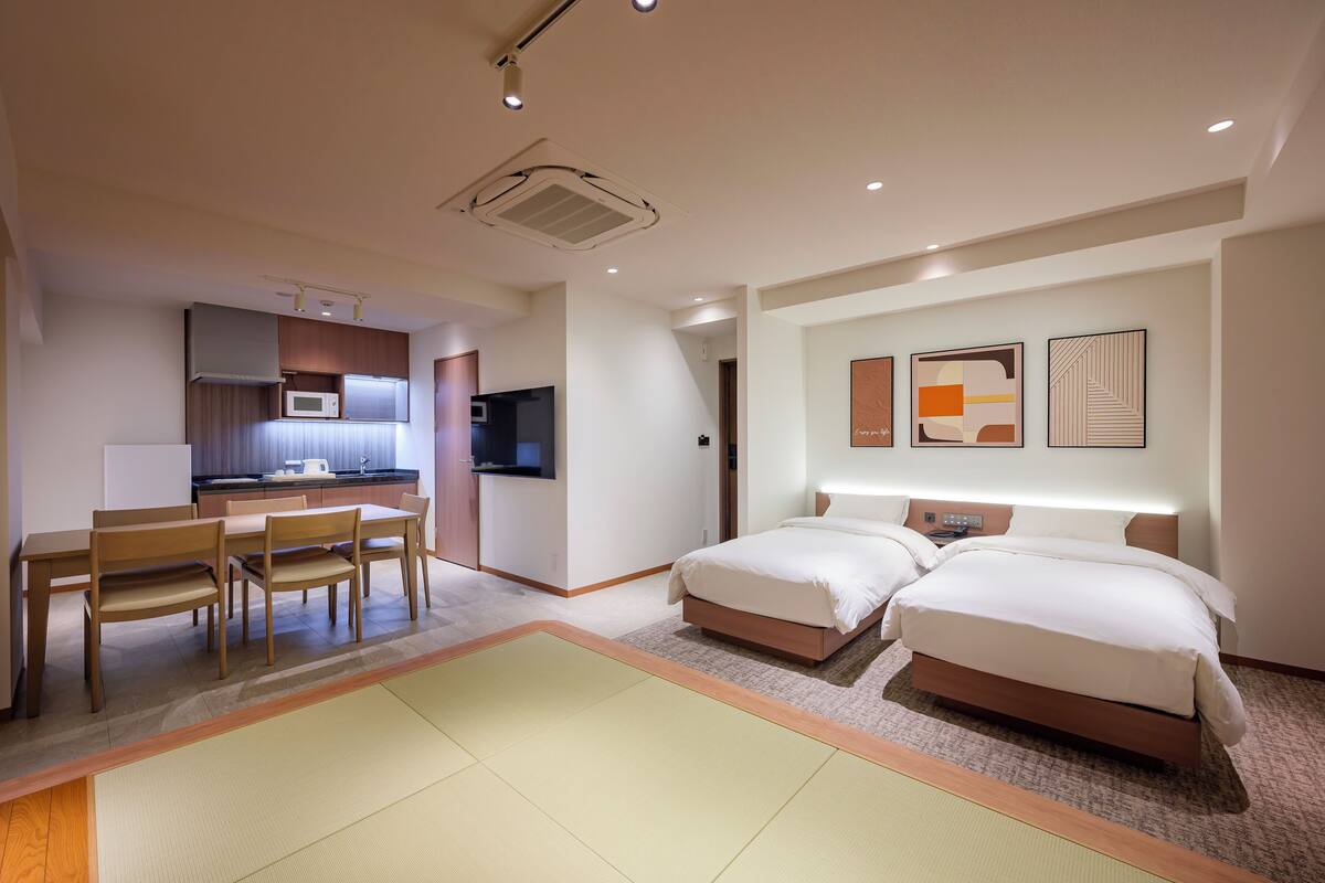 Une chambre d’hôtel comprenant un lit, une table de chevet et un petit bureau avec une chaise. Elle dispose également d’une kitchenette équipée d’un four à micro-ondes et d’un évier.