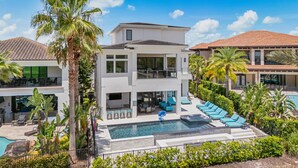 A modern two-story house with a swimming pool, surrounded by palm trees and other greenery.