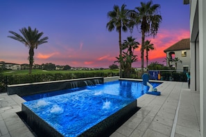 Una piscina con cascada, rodeada de palmeras y vistas a un campo de golf al atardecer.