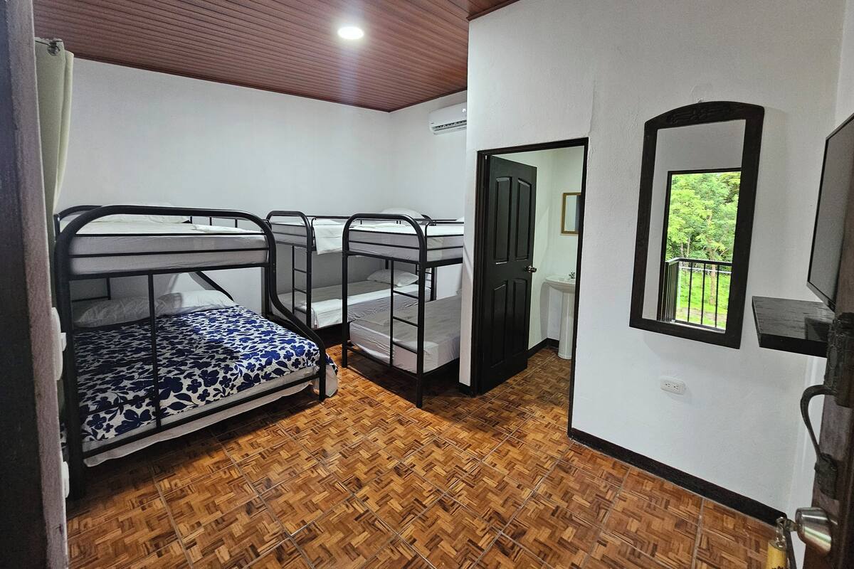 A room with four bunk beds, a wooden floor, a white wall, a door leading to a bathroom, and a window with a view of greenery.