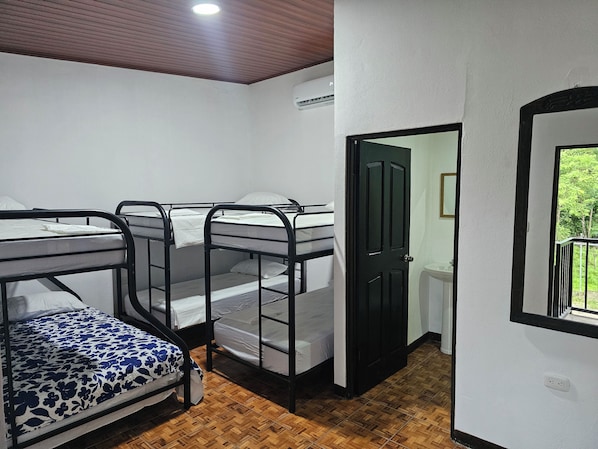 A dormitory room with bunk beds, a wooden floor, a white wall, a door, and a window.
