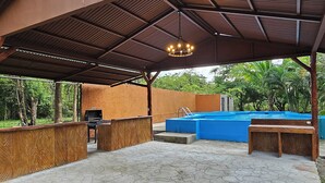 Área de piscina externa com um alpendre coberto, churrasqueira e deck de madeira.