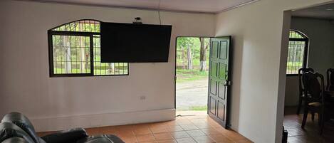 A spacious living room with a large flat-screen TV mounted on the wall, a leather sofa, and a view of the outdoors through a window.