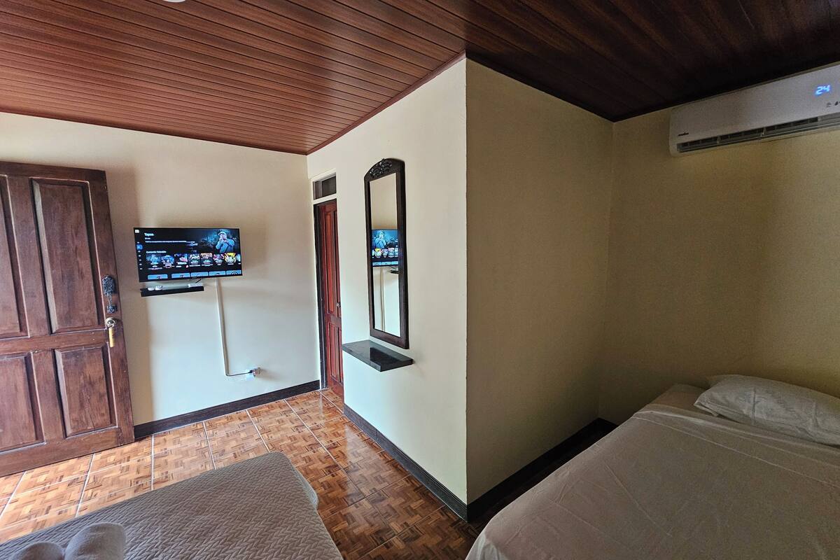 A hotel room with a wooden ceiling, a television mounted on the wall, a bed with white bedding, and a wooden door.