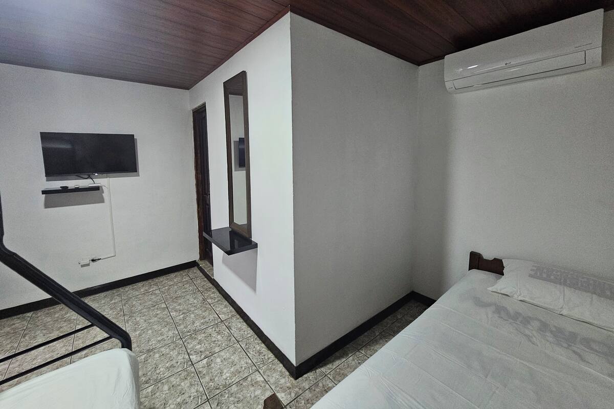 A hotel room with a bed, a TV mounted on the wall, a mirror, and an air conditioning unit.
